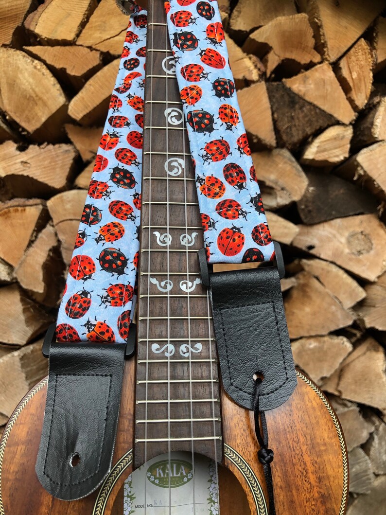 May include: A ukulele with a black leather strap and a blue and white ladybug patterned strap. The ukulele has a wooden body and silver-coloured tuning pegs. The ukulele is labelled "KALA" on the body.