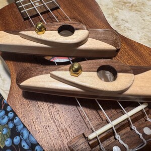 Pode incluir: Close-up de um ukulele com corpo de madeira e detalhes em madeira clara. A ponte e a sela são visíveis, juntamente com as cordas e as cravelhas. Uma correia estampada em azul e branco é visível à esquerda.