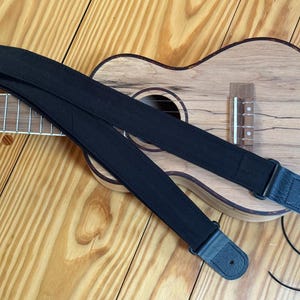 Może przedstawiać: Czarny pasek do ukulele ze skórzanymi końcami spoczywa na drewnianym ukulele. Pasek wykonany jest z wytrzymałego, tkanego materiału. Ukulele ma naturalne drewniane wykończenie i jest umieszczone na drewnianej powierzchni.