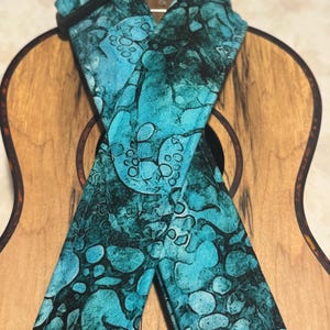 May include: A teal and black ukulele strap with a unique pattern. The strap is made of fabric and features a repeating design of organic shapes. The strap is shown over a wooden ukulele.