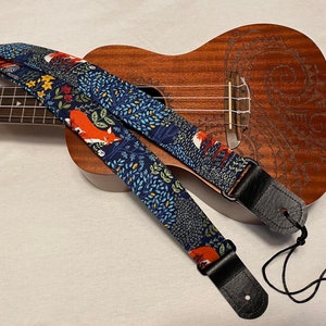 May include: A ukulele strap with a navy blue background featuring a repeating pattern of orange foxes, green foliage, and yellow flowers. The strap has black leather ends and is resting on a wooden ukulele.