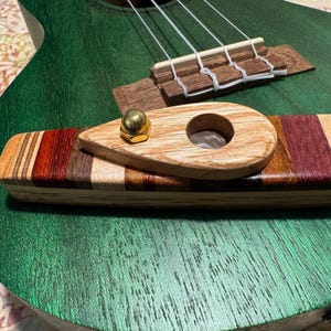 Pode incluir: Um close-up de um ukulele verde com uma ponte e cordas de madeira. Um capo de ukulele de madeira com uma porca dourada está em primeiro plano. O capo é feito de várias tonalidades de madeira e tem um buraco no centro.