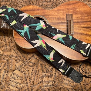 Może przedstawiać: Czarny pasek do gitary z wzorem kolibrów. Pasek jest wykonany z tkaniny i ma skórzany koniec. Pasek leży na brązowej powierzchni z nadrukowanymi nutami.