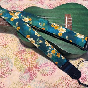 Może przedstawiać: Turkusowy pasek do ukulele z kwiatowym wzorem z białych i żółtych kwiatów oraz zielonych liści. Pasek leży na zielonym ukulele. Pasek ma czarne skórzane końce i czarny sznurek do mocowania.
