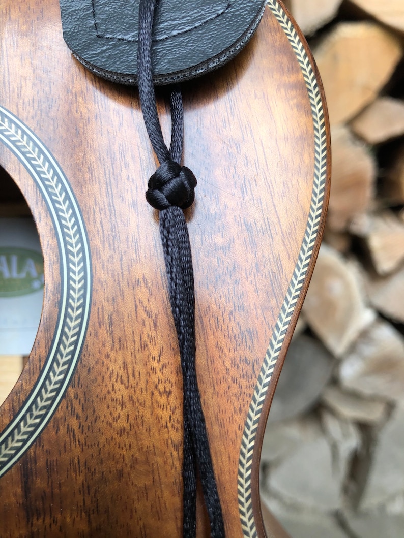 May include: A close-up of a ukulele with a black strap attached to the body. The ukulele is made of wood and has a gold-coloured inlay around the sound hole.