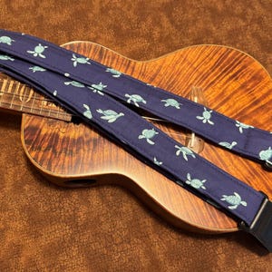 May include: A blue guitar strap with a pattern of green sea turtles. The strap is attached to a ukulele with a brown wooden body.