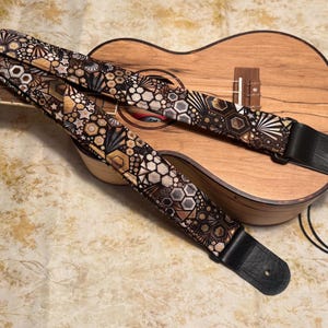 May include: A patterned ukulele strap with a geometric design in black, brown, and tan, resting on a wooden ukulele. The strap has black leather ends and a black cord for attachment. The ukulele has a natural wood finish.