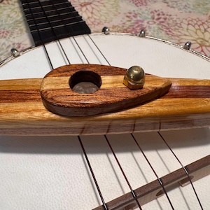 Pode incluir: Close-up da ponte e do esticador de um banjo. A ponte é feita de madeira clara e escura, com uma porca de latão. As cordas são visíveis, juntamente com a cabeça branca e a borda de metal do banjo.