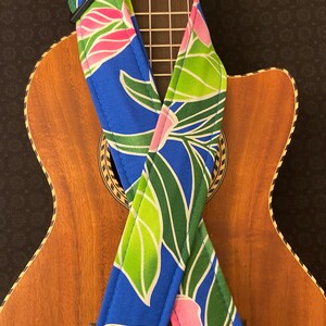 May include: A blue ukulele strap with a tropical floral pattern featuring green leaves and pink flowers. The strap is shown on a wooden ukulele.