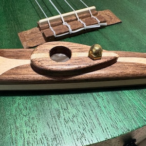 Pode incluir: Close-up da ponte e da cobertura do orifício sonoro de um ukulele. A ponte e a cobertura são feitas de madeira escura e clara, com um botão de latão na cobertura. O corpo do ukulele é verde.