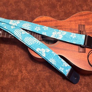 May include: A blue ukulele strap with a white floral pattern. The strap is attached to a brown ukulele.