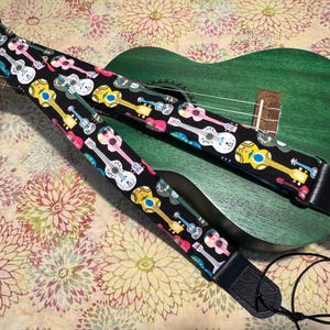 Może przedstawiać: Czarny pasek do ukulele z kolorowym wzorem gitary, spoczywający na zielonym ukulele. Pasek przedstawia różne wzory ukulele w kolorze białym, żółtym, różowym i niebieskim. Tło ma kwiatowy wzór.