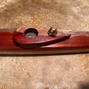 Pode incluir: Um cachimbo de madeira artesanal com acabamento castanho-avermelhado escuro e base de madeira mais clara. O cachimbo tem uma tigela circular e um detalhe de porca de latão. O design é retangular com bordas arredondadas.