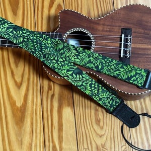 May include: A ukulele strap featuring a vibrant green and black leaf pattern, constructed with fabric and black leather ends. The strap includes a black plastic adjustment buckle and a thin black cord for attachment to the instrument's headstock.