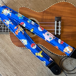 Puede incluir: Una correa de ukelele azul con un diseño de personaje de Pokémon. La correa presenta varios personajes de Pokémon, incluidos Pikachu, Eevee y Jigglypuff, sobre un fondo azul con Poké Balls.