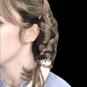 Puede incluir: Vista lateral de la cabeza y el hombro de una persona, con cabello castaño peinado en trenza y cola de caballo. El cabello está asegurado con una goma de pelo en espiral marrón. La persona lleva varios pendientes y un gorro blanco de ganchillo.