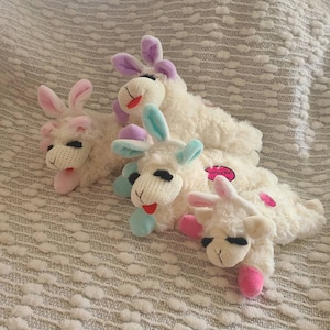 May include: Four plush white stuffed animals shaped like lambs with different colored ears. The lambs are lying down on a white textured surface. The ears are pink, purple, and blue.