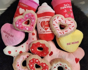 May include: A collection of Valentine's Day themed dog toys. The toys are shaped like donuts, hearts, and coffee cups. The toys are pink, red, and yellow with various embroidered details. The toys feature phrases like "Droolin' 4 U", "I love you", "Be Mine", "You're Pawfect", "#PuppyLove", and "Lippy Love".