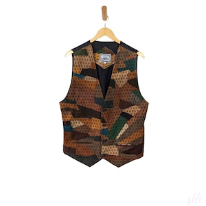Könnte beinhalten: Eine Patchwork-Weste in Braun-, Grün- und Blautönen hängt an einem Holzbügel. Die Weste weist eine Vielzahl geometrischer Formen und Muster auf. Sie hat ein schwarzes Futter und eine Knopfleiste. Die Weste ist ein einzigartiges Kleidungsstück.