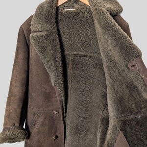 May include: A brown suede coat with a shearling lining. The coat has a wide collar, button closures, and fur trim on the cuffs. The interior is a darker shade of brown than the exterior. The coat is hanging on a wooden hanger.