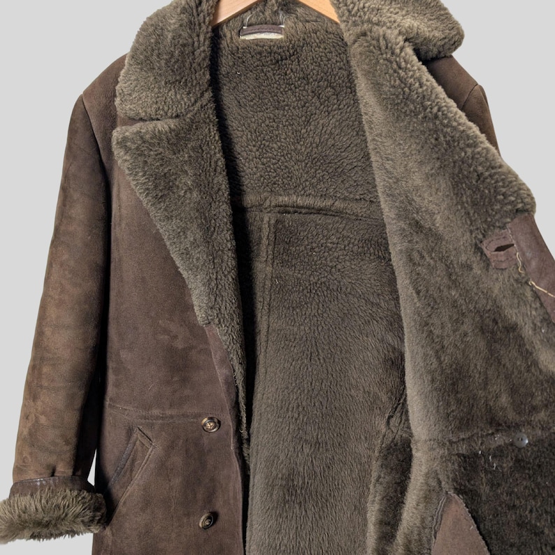 May include: A brown suede coat with a shearling lining. The coat has a wide collar, button closures, and fur trim on the cuffs. The interior is a darker shade of brown than the exterior. The coat is hanging on a wooden hanger.