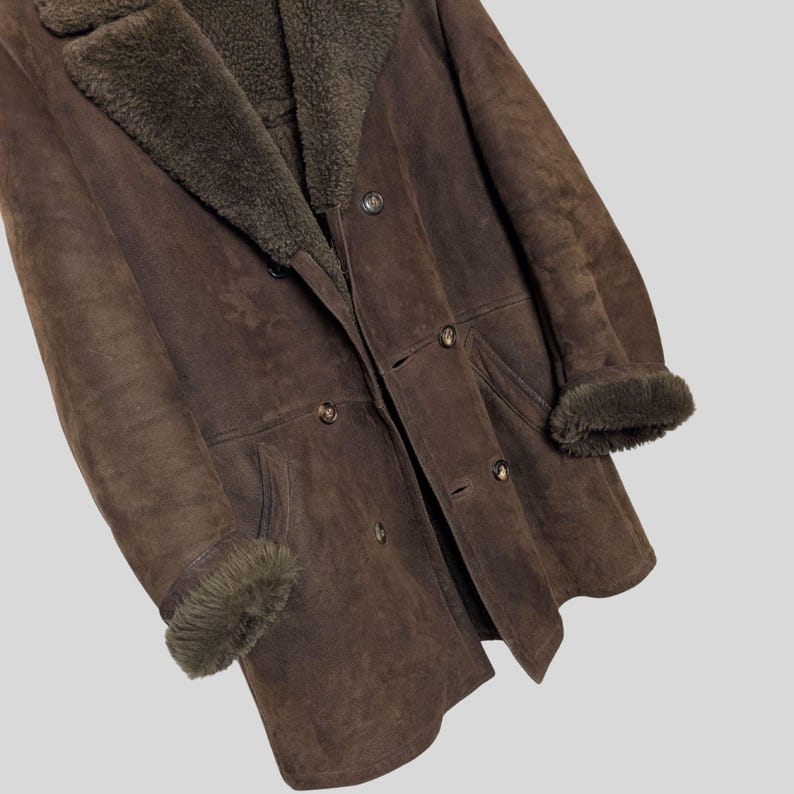 May include: A brown shearling coat with a double-breasted design. The coat features a plush, dark brown shearling lining and collar, with matching cuffs. The outer layer is a darker brown suede-like material, and the coat has several buttons.