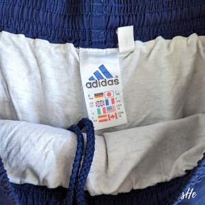 以下が含まれることがあります： 白いタグが付いたネイビーブルーのadidasのドローストリング付きアスレチックショーツ。タグには、インチとセンチメートルでサイズ情報が記載されています。タグには、adidasのロゴと「adidas」の文字も記載されています。
