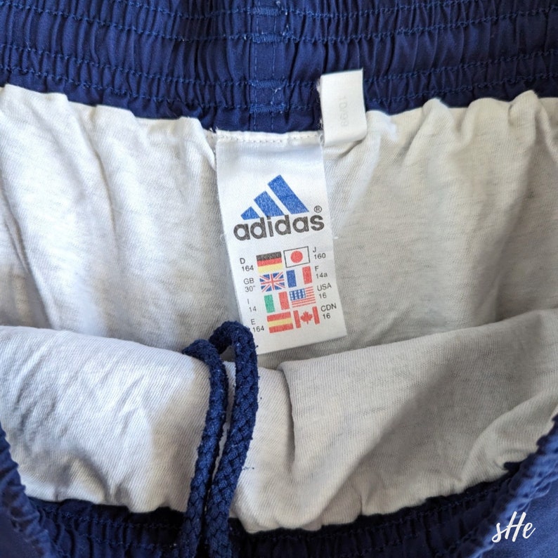 以下が含まれることがあります： 白いタグが付いたネイビーブルーのadidasのドローストリング付きアスレチックショーツ。タグには、インチとセンチメートルでサイズ情報が記載されています。タグには、adidasのロゴと「adidas」の文字も記載されています。
