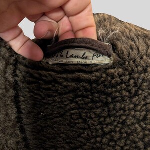 May include: Close-up of a brown, textured garment, possibly a coat or jacket, with a label that reads "100% Lambs Fur". The label also states "Coordinated and Made in France". The garment appears to be made of a soft, plush material.