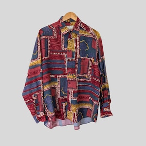 Könnte beinhalten: Langärmliges Hemd mit farbenfrohem geometrischem Muster. Das Hemd hat eine Mischung aus roten, blauen, gelben und weißen Formen. Das Hemd hängt an einem Holzbügel.