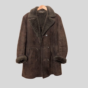 May include: A brown, double-breasted coat with a shearling collar and cuffs. The coat is hanging on a wooden hanger. The coat has a classic design with a warm, textured appearance.