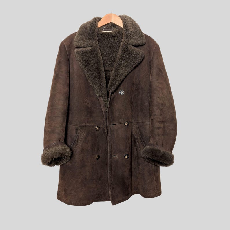 May include: A brown, double-breasted coat with a shearling collar and cuffs. The coat is hanging on a wooden hanger. The coat has a classic design with a warm, textured appearance.