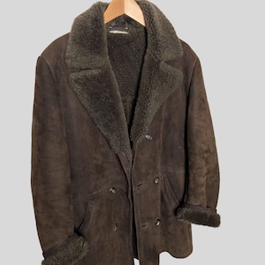 May include: A brown suede coat with a shearling collar and lining. The double-breasted coat has two rows of buttons and a classic design. The cuffs are trimmed with shearling. The coat is hanging on a wooden hanger.