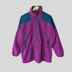 Könnte beinhalten: Eine lila Fleecejacke mit tealfarbenen Schulterpartien und tealfarbenen Reißverschlüssen. Die Jacke hat einen hohen Kragen, einen durchgehenden Reißverschluss und zwei Reißverschlusstaschen. Ein Kordelzug verengt die Taille. Das Columbia-Logo ist sichtbar.