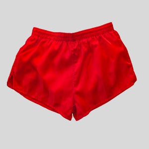 May include: Bright red athletic shorts with an elastic waistband. The shorts are made of a lightweight, shiny material. These running shorts are designed for comfort and ease of movement.