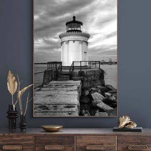 May include: Black and white photograph of a lighthouse on a rocky shore. The lighthouse is tall and white with a black roof and a black lantern. The shore is rocky and the water is calm.