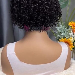 May include: A short, curly black wig styled on a mannequin. The wig is densely packed with tight curls, offering a voluminous look. The mannequin is wearing a white top, and the background includes flowers and greenery.