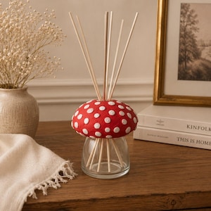 May include: A reed diffuser featuring a clear glass base and a red mushroom-shaped cap with white polka dots. Several thin, light-coloured wooden reeds extend upwards from the centre of the mushroom cap, creating a functional and decorative home accessory.
