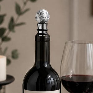 May include: A bottle stopper featuring a spherical white and grey marbled stone top attached to a silver-toned metal base with black rubber rings, inserted into the neck of a dark glass bottle filled with a dark liquid.
