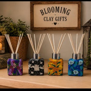 May include: Four square glass reed diffusers with silver collars and wooden reeds. The bottles feature distinct patterns: purple and green floral, black and white geometric, yellow and orange floral, and blue and white floral designs.