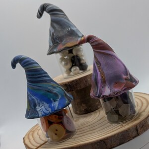 May include: Three decorative glass jars with whimsical, swirled-color gnome hats. The jars are filled with buttons, pearls, and stones. The hats are in shades of blue, purple, and black, with gold accents. The jars sit on a wooden slice.