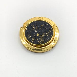 May include: A circular gold-tone metal bag hanger featuring a textured black centre inlay with scattered gold-coloured flecks. The device has a polished metallic frame with a hinged design, intended to rest on a flat surface to support a bag.