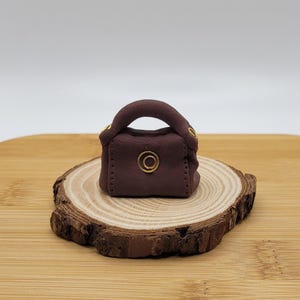 May include: A miniature brown handbag with a rounded handle and gold-colored accents. The bag features a circular gold detail on the front and is placed on a wooden slice. The background is a neutral white.
