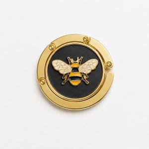 May include: A circular gold-toned metal bag hanger featuring a central black disc inlay. The center of the disc displays a detailed bee motif with yellow and black striped body segments and cream-colored wings, all rendered in a smooth enamel finish.