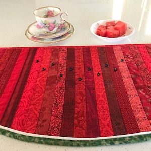 May include: A red and green watermelon-shaped table runner with a white border. The runner is made of fabric strips sewn together to create a striped pattern. There are black beads sewn onto the runner, resembling watermelon seeds.