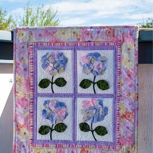 May include: A purple and pink floral quilt with four squares featuring a 3D flower design. The quilt is hanging on a white wooden fence against a blue sky.