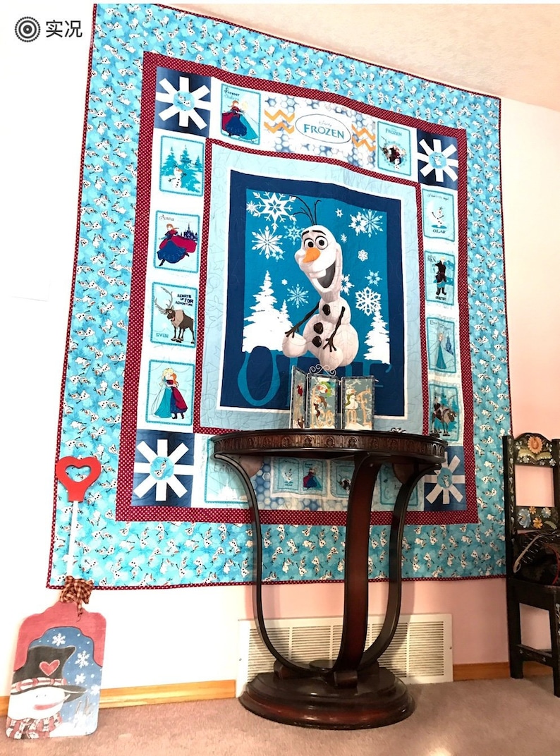 Puede incluir: Una colcha azul y blanca con tem&aacute;tica de Frozen con Olaf el mu&ntilde;eco de nieve. La colcha est&aacute; colgada en una pared con una mesa y una silla de madera en primer plano.