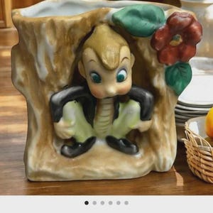 May include: A vintage ceramic planter shaped like a tree trunk, featuring a whimsical elf-like figure. The planter is painted in earthy tones with a red flower and green leaves. The elf has a black jacket and green trousers.