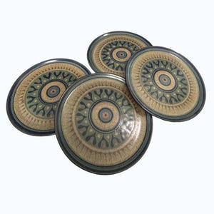 May include: A set of four ceramic plates with a decorative design. The plates have a beige background with a repeating pattern in green, blue, and brown. The plates have a dark blue rim.