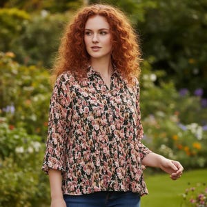 May include: A floral print blouse with a collared neckline and three-quarter sleeves. The blouse features a pattern of pink roses on a black background. The model is wearing blue jeans. The background is a garden.